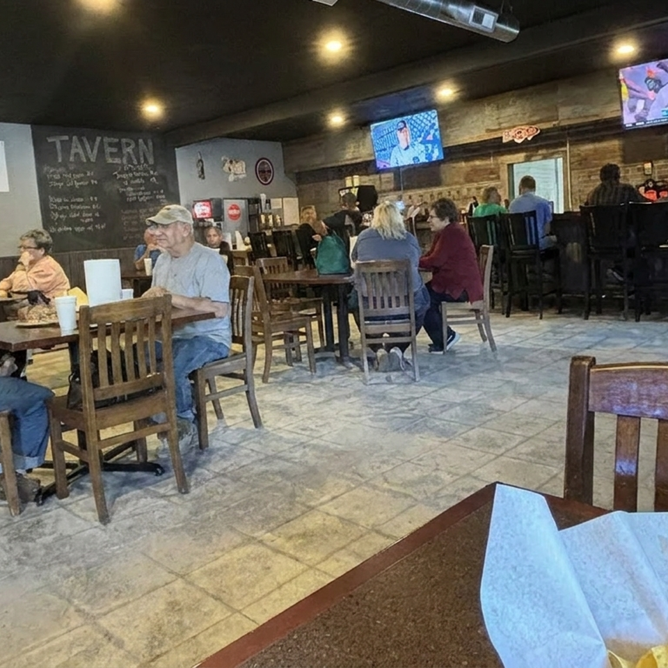 Busy rustic tavern dining room with patrons at wooden tables, bar seating, televisions, chalkboard menus, and a server behind the counter.
